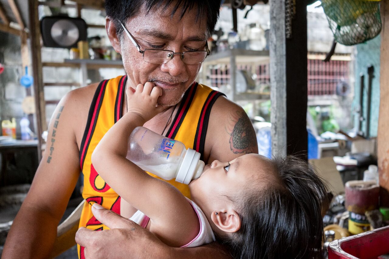 Orlando feeds his granddaughter at his home in Tarlac, Philippines. Orlando's wife went to work as a domestic worker in Hong Kong over 20 years ago and has yet to return. It is hard to establish trust in the relationship. “She always asks where I am. So I started this Sari Sari [small convenience shop] last year, just to show her I am busy at home.”