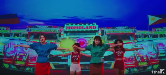 The girls of Momoland dancing in front of a backdrop where images of jeepneys and two Philippine flags were projected. Screenshot from the video teaser.