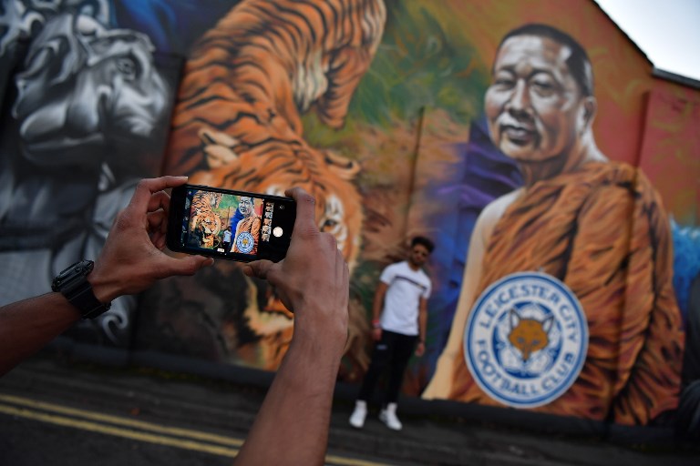 A man poses for a photograph against a mural showing Leicester City Football Club’s Thai chairman Vichai Srivaddhanaprabha near De Montfort University in Leicester, eastern England, on October 28, 2018 after a helicopter belonging to Srivaddhanaprabha crashed outside Leicester City Football Club’s King Power Stadium the night before. Ben Stansall / AFP
