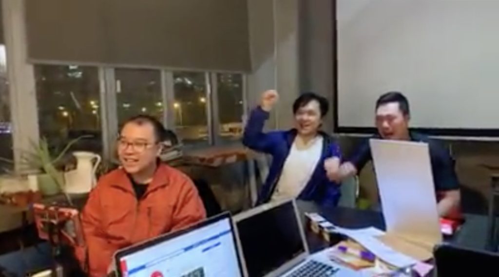 Colleagues of the two men cheer on two men attempting to eat an entire tin of Garden Bakery cookies. Screengrab via Facebook video/100most.