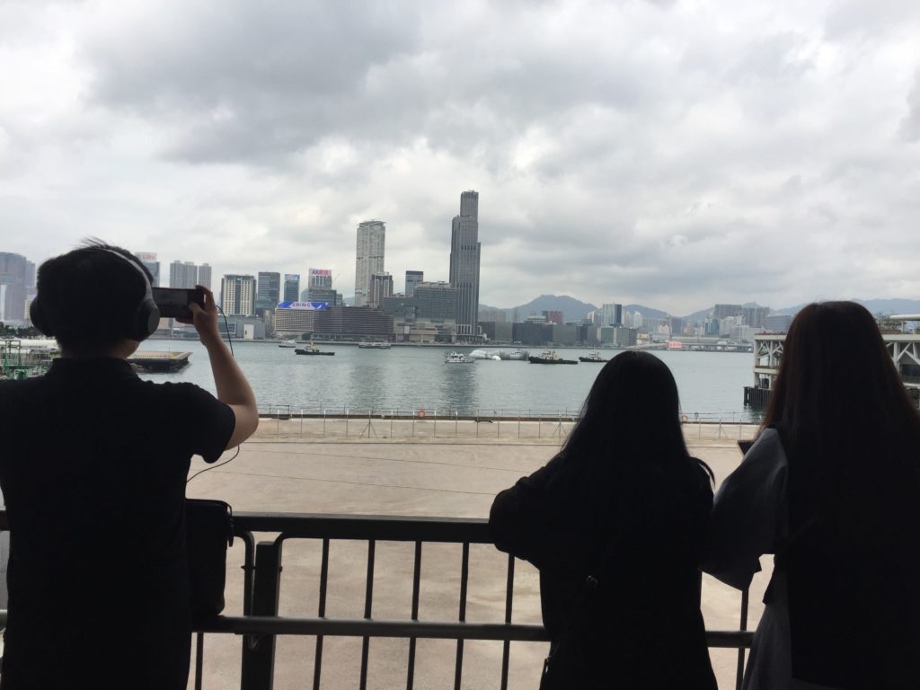 Onlookers snap photos of the Kaws installation as its towed past the Wan Chai ferry pier today. Photo by Stuart White.