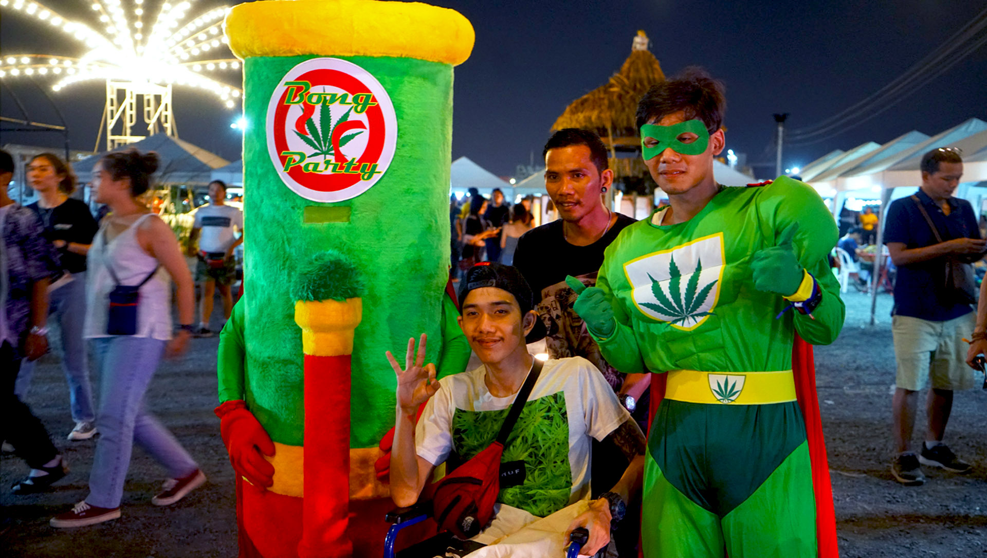 A young man in a wheelchair poses with a human bong mascot and Super Weedman on Saturday at the Runway 3119 Night Market in Bangkok.