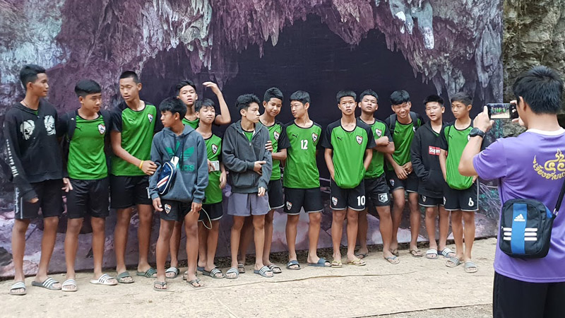 Young footballers who said they were ‘dogs’ not ‘wild boars,’ pose for a photo in January 2019 before a backdrop erected outside the Tham Luang cave entrance in Chiang Rai province. Photo: Coconuts Media