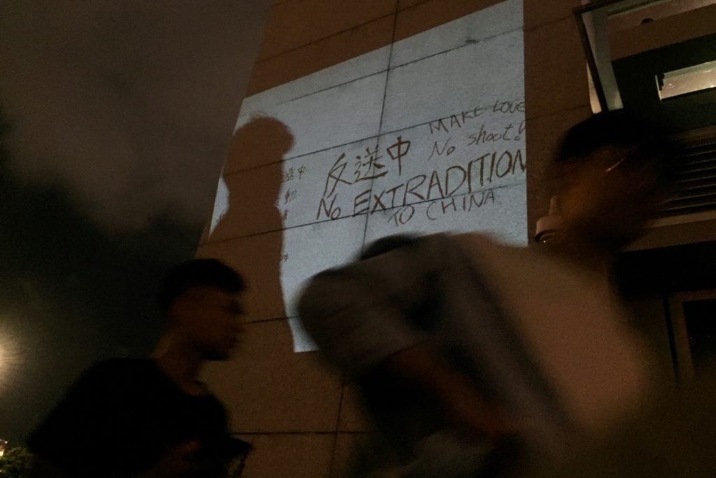 A protester uses a mini-projector to display a sign drawn by a fellow demonstrator who fell to his death last week on a wall near tonight's protest. Photo by Stuart White.