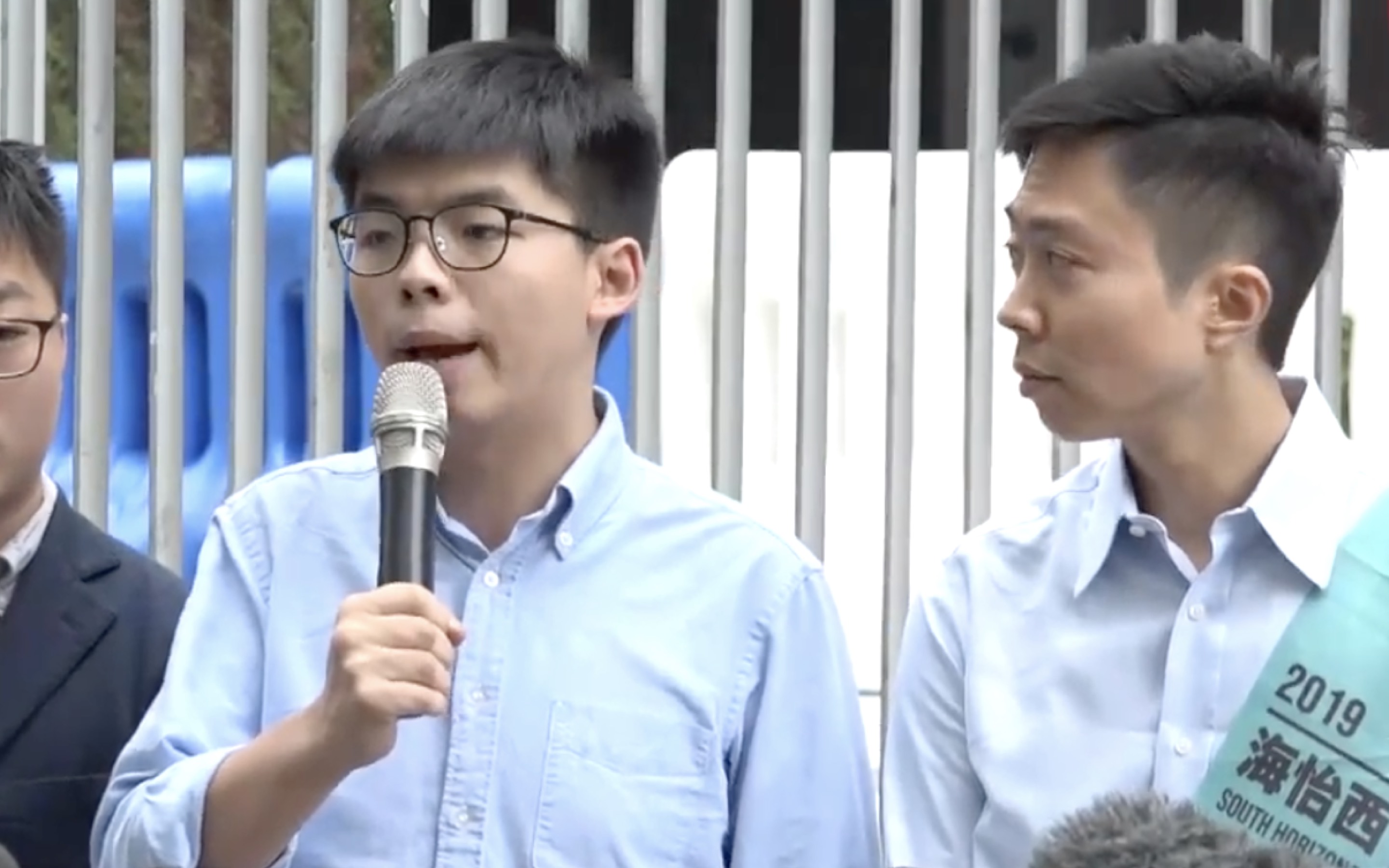 Joshua Wong (left) speaks to reporters hours after a returning officer barred him from running in the upcoming district council elections on Nov. 24. He urged voters to support the pro-democracy side’s ‘plan B candidate’ Kelvin Lam (right). Screengrab via YouTube/Apple Daily.