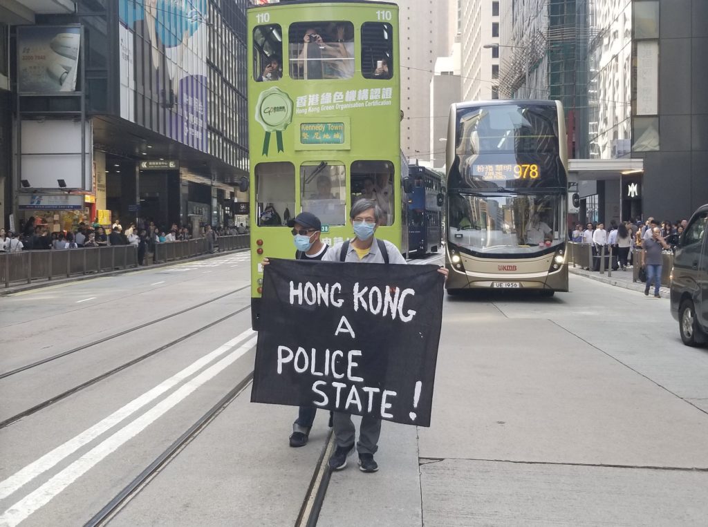 Man holding a banner reading 'Hong Kong a police state!' stands in front of a tram. Photo by Vicky Wong.