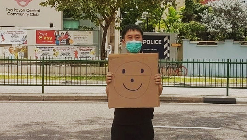 Social worker and civil rights advocate Jolovan Wham poses with smiley face placard in Toa Payoh in a photo that has been cropped and brightened. Photo: @JWham/Instagram