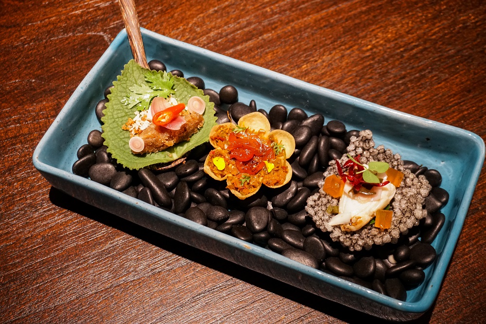 A tray of three bite-sized pieces, from right to left: crab salad on a squid ink-soaked sago cracker; a tart of smoked catfish, chili paste and herbal relish; and Indian borage leaf wrapping. Photo: Coconuts