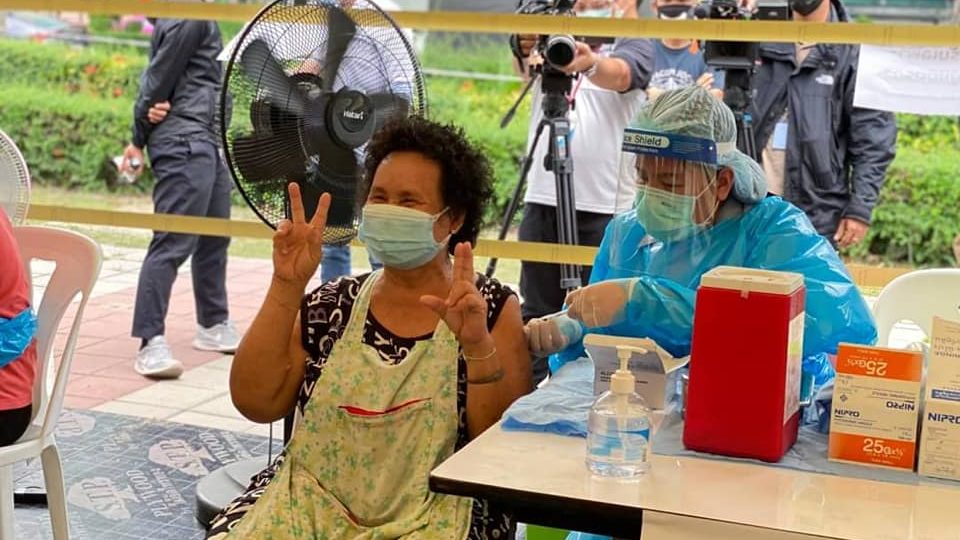 
A Khlong Toei resident raises two fingers for a camera while being vaccinated. Photo: Khlong Toei District Office
