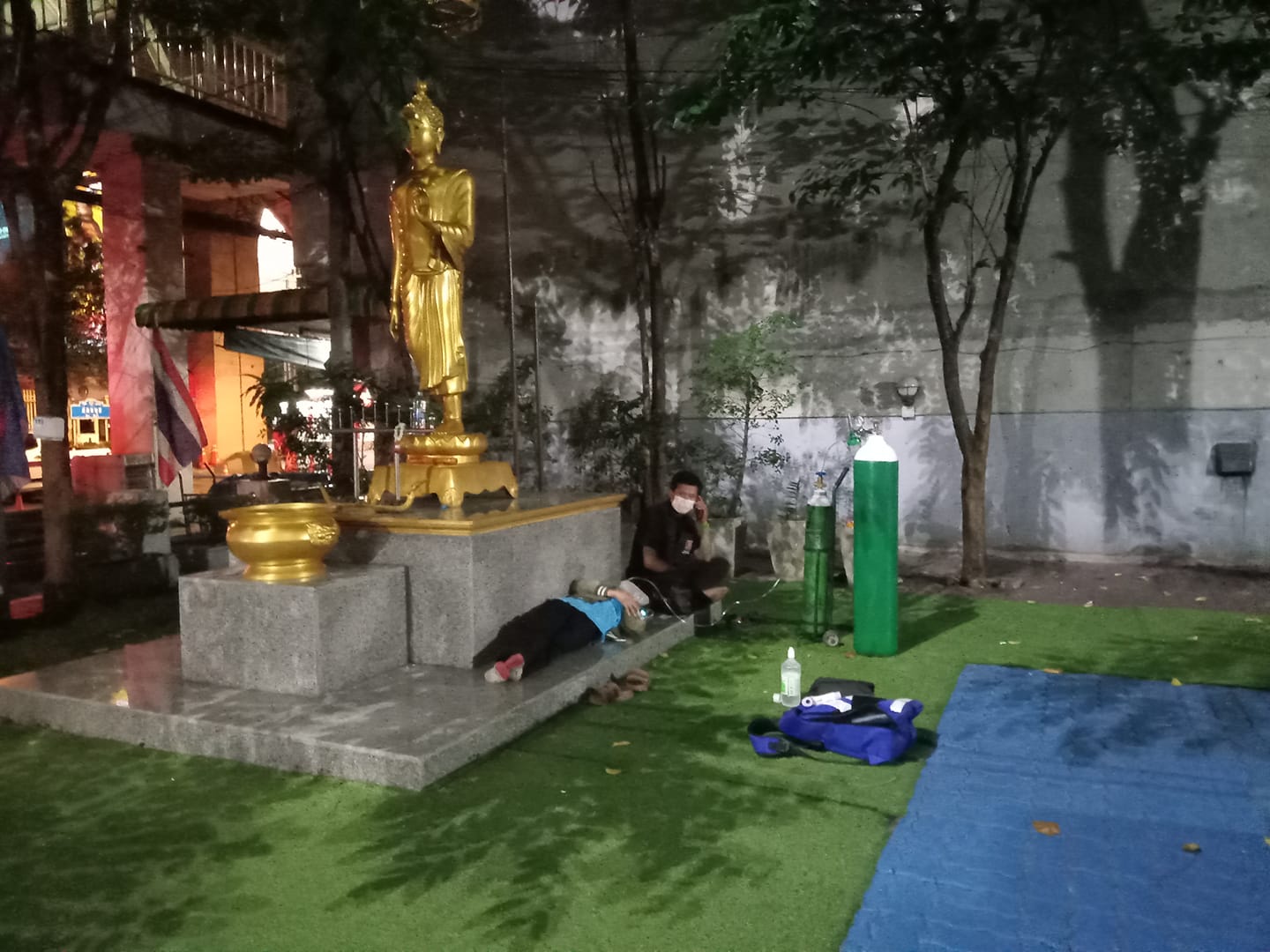 Waiting for a hospital bed, a woman receives oxygen supply on Tuesday night at the Phra Khanong Police Station with her husband sitting next to her. Photo: Dekpichai Khontokkung / Facebook
